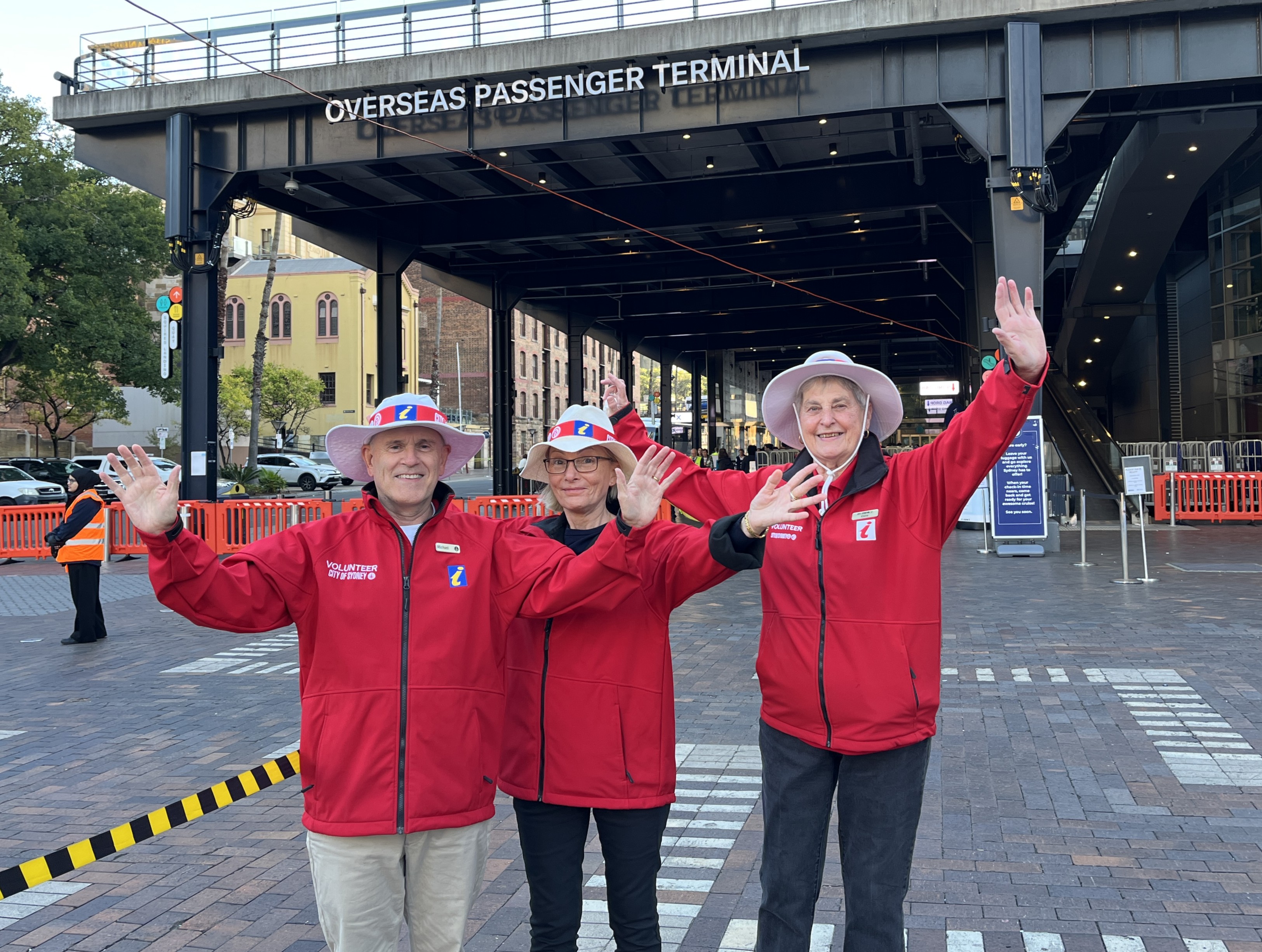 Three smiling Cruise Ambassadors wearing bright red jackets with their hands raised in front of Overseas Passenger Terminal