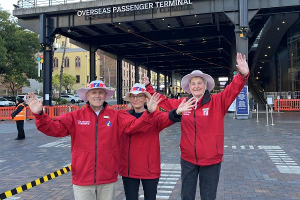 Three smiling Cruise Ambassadors wearing bright red jackets with their hands raised in front of Overseas Passenger Terminal