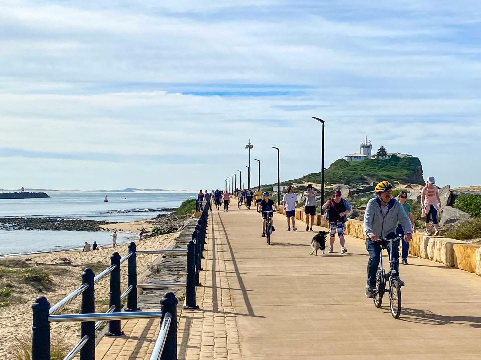 Members of the public walking and cycling along Macquarie Pier with Nobbys in the distance