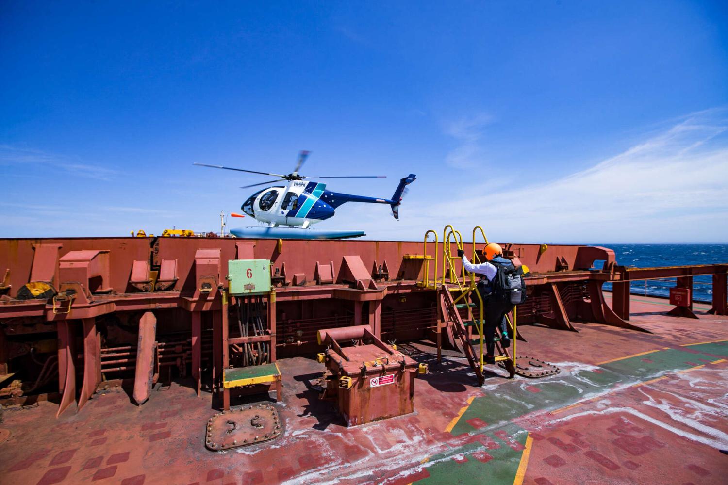 On top of a large ship, a pilot climbs a ladder to a helicopter that is hovering nearby