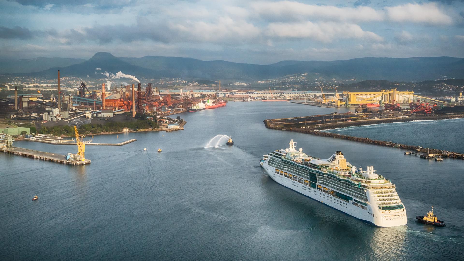 A cruise ship enters Port Kembla escorted by two tugs with Wollongong in the distance