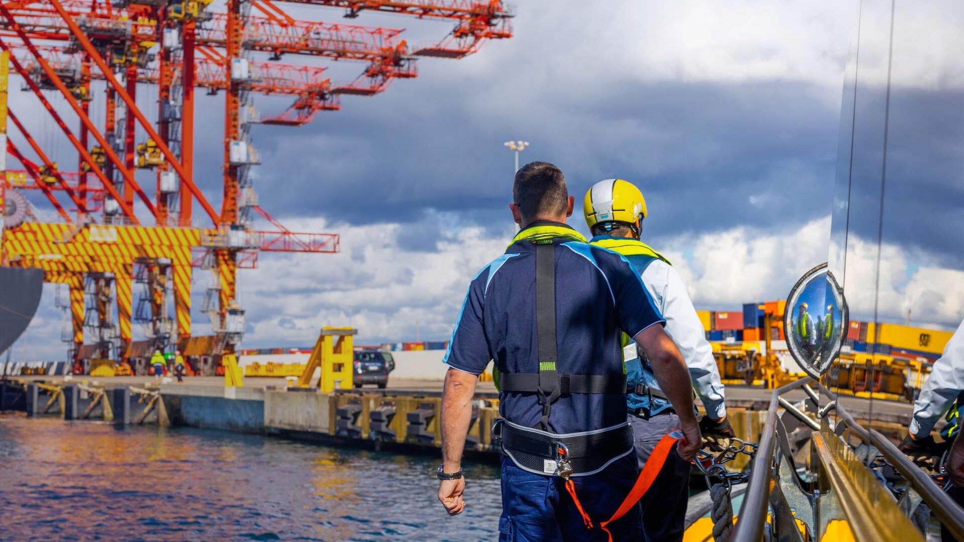 Two Port Authority employees walk alongside a vessel on the water at Port Botany with cranes in distance