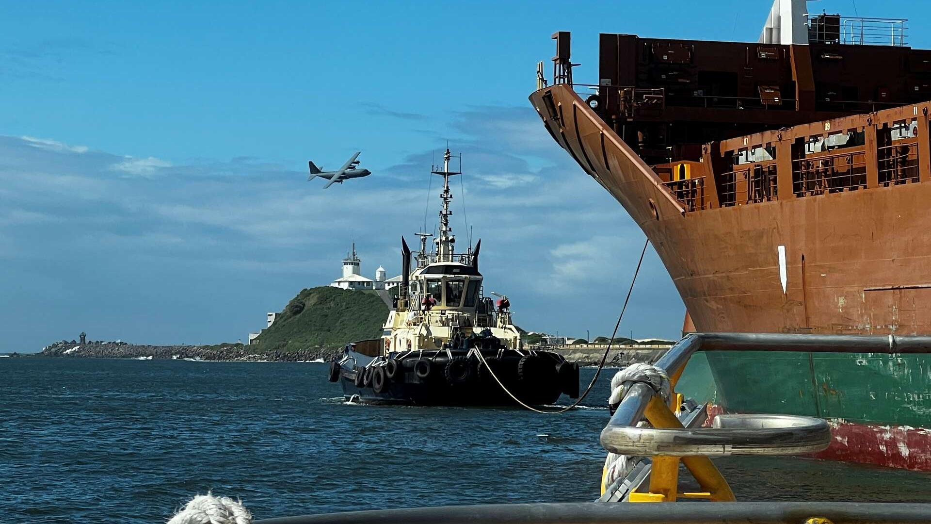 A plane flies over Nobbys Headland with tug on the water